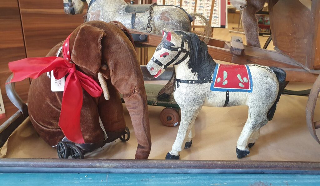 A soft brown velvet elephant on wheels with a big red ribbon and a model horse with painted saddle and reins sit looking at each other. Both are vintage and well loved.