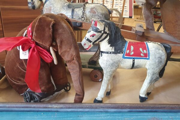 A soft brown velvet elephant on wheels with a big red ribbon and a model horse with painted saddle and reins sit looking at each other. Both are vintage and well loved.