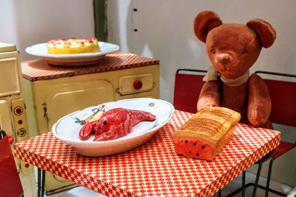 a small brown velvet traditional teddy bear sits at a small metal table in a dollhouse kitchen diorama. on the table in front of him is a miniature loaf of bread and a china dish with a model lobster.